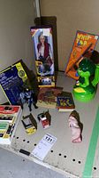 Overview of lot on a shelf showing boxed Cherokee Barbie, Green Giants Niblet flashlight, Shirley Temple book, Batman action figure, two toy vehicles, small books and baby figure, all arranged for view.