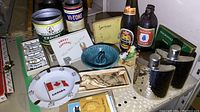 Zoomed-out view of a shelf with two stainless steel hip flasks, ceramic and glass ashtrays, vintage tobacco packaging, liquor bottle tags, and other barware-related items.