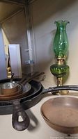 Photo showing small copper clad frypans stacked inside cast iron frypans with a green glass hurricane lamp in the background.