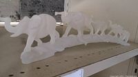 Side view of frosted elephant figurine set on shelf, showing the varying sizes of the 5 elephants walking in a line on a curved base.