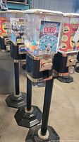 Photo showing multiple metal and plastic double gumball machines on black pedestal stands, featuring colorful labels for items dispensed like 'Glo-in-The-Dark Aliens' and 'Fuzzy Faces Moustaches'.
