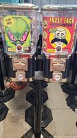 Front view of two standing metal and plastic gumball machines side by side with colorful graphics and coin slots visible.