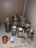 Collection of 14 assorted steins including ceramic, metal-topped and glass mini steins of various sizes and decorative styles.