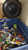 Large blue bin filled with various loose small toy cars and trucks, showing many different colors and styles. Super Rally tire-shaped carrying case visible behind bin.