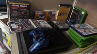 Black PS4 Slim console on shelf with controller on top, game cases behind and to side