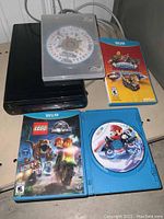 Black Wii U console with cords and four game discs in cases, showing LEGO Jurassic World, Mario Kart 8, Skylanders SuperChargers and an unlabeled disc in a clear plastic case.