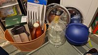 Kitchen items grouped including orange Char Buster cutting set box, copper-colored pump carafe, knife set in packaging, clear glass beverage carafe, and blue plastic container with lid.