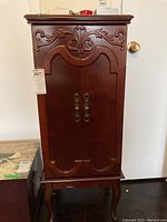 Front view of wooden jewelry armoire with carved floral patterns and brass handles on double doors.