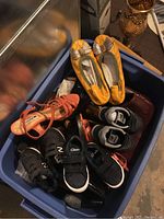 Blue bin containing ladies and toddlers shoes including yellow flats with silver bows, black Clarks sneakers, black New Balance toddler sneakers, orange strappy sandals, and other footwear.