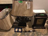 Photo showing one table lamp with beige fabric lampshade and ornate dark base, wood framed mantel clock next to it, two small blue items in front, set on concrete floor.