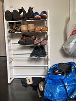 Wide view of white metal shoe rack holding various ladies shoes including boots, sandals, flats, and sneakers. Blue IKEA bag with shoes on the floor beside rack.