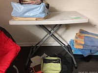 Side view of folding utility table with folded metal legs and white textured plastic top. Table is holding several bags and fabric items.