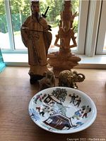 Photo of the six items: two Asian figurines, two elephant figurines, and one decorated ceramic dish on a wooden surface with window background.