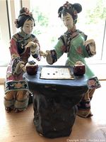 Front view of two detailed Asian styled porcelain statues of women playing a board game on a black carved pedestal table