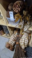 Photo of large natural pinecones, faux autumn leaves, a small broom, and autumn scarecrow dolls on shelving unit.