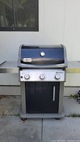 Front-facing view of Weber Spirit gas grill. Black lid with temperature gauge and silver Weber logo. Three control knobs and side shelves visible.