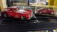 Side view showing 1954 Mercedes Benz Gullwing SL300 model alongside 1948 Tucker in display case, both red.