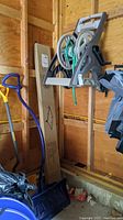 Photo showing hose reel mounted on wheels with garden hose wound around it, and two snow shovels (yellow and blue) standing beside it inside a wooden shed or garage.