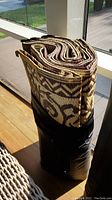 Rolled indoor/outdoor rug with a brown and cream geometric and leaf pattern, secured in a black carrying case or sleeve, placed upright on a wooden floor near a window.