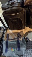 Photo of gray tote bag with navy blue handles and colorful embroidered design, and a black suitcase on casters with front zippered pocket