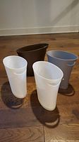 Four plastic wastepaper baskets in white, brown, and gray with oval shape and handles, shown on wood floor.