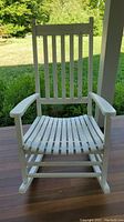 Front view of the white wooden outdoor rocking chair placed on a wooden porch with greenery in the background showing vertical slats on backrest and slatted seat.