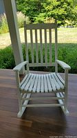 White wooden outdoor rocking chair with vertical slat back and curved slat seat, showing wear and weathering on the paint. The chair is positioned on a wood deck with green outdoor scenery.