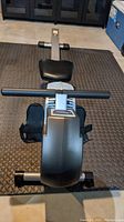 Top view of black cushioned seat, handlebar with foam grip, and digital console on front of silver frame of Stamina ATS 1399 rowing machine on black textured floor mat in basement.