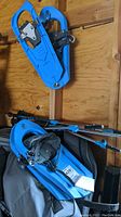 Six pairs of blue Yukon Charlies snowshoes with attached black bindings and cleats, stacked together inside carrying bags on a wooden background.