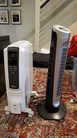 Both DeLonghi white oil-filled heater on wheels and Holmes black tower fan heater side by side on carpeted floor.