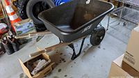 Black ACE plastic wheelbarrow with two wheels, steel frame and legs. Weathered condition with visible small crack and oxidation. Located inside a garage with various items in background.