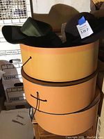 Two hats placed atop stacked large round hat boxes with black drawstrings. Hats include a black hat with a blue price tag and a dark green hat with a wide brim.