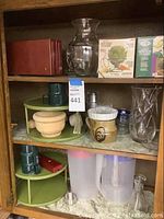 Shelf with assorted glassware, plastic rotating organizers, artichoke strainers, pitchers, and ceramic cup.