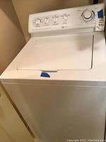 Front and top view of the Maytag washing machine showing the white lid and some blue tape on the corner.