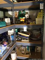 Utility shelf with four shelves holding various household and office items such as glass bowls, boxes, jars, and packaged goods in a storage or utility room environment.