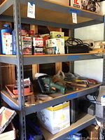 Photo of a gray-blue metal utility shelf with four shelves, showing various items stored including charcoal lighter fluid and tools. The shelf is empty for sale.