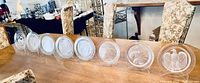 Eight clear Lalique crystal plates with etched animal and nature motifs, displayed on plate stands on a wooden table with floral upholstered chairs in the background.