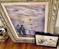 Large framed painting with two men looking out over ocean at sunset, birds flying, with smaller framed painting and other small artwork leaning against it