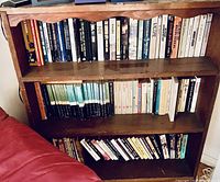 Wooden bookshelf with three shelves filled with books of various genres and sizes. Books are in good condition with visible spines showing titles and authors.