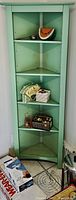 Full perspective view of green corner wooden stand arranged with assorted coasters, baskets, and other small kitchen items on shelves, visible against white wall and tiled floor.