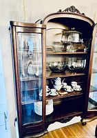 Wide view of vintage wooden display case with glass shelves, housing all dishware items including clear bowls, plates, and china sets.