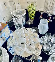 Wide view showing various glass and plastic drinkware including punch bowl, martini glasses, and pitcher among other glassware on a table.