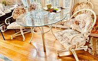 Photo of the 48" round glass dining table with cream metal base and four fabric rolling chairs arranged around it on a hardwood floor.