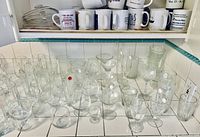 Array of assorted clear drinking glasses, stemmed glasses, goblets, tumblers and six white ceramic coffee mugs with printed logos.