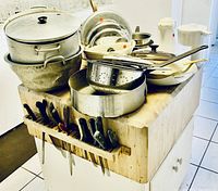 A kitchen island or cabinet with a large wooden butcher block cutting board holding multiple stainless steel roasting pans, pots, colander, pans, lids, and a coffee pot, along with a white Brita pitcher. Visible kitchen knives are inserted in slots on the cutting board.