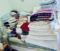 First image showing stacks of folded sheets, comforters, blankets, and towels piled on a bed against the wall.