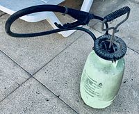 Close-up of a portable pump sprayer with black handle, trigger and hose on paved surface.