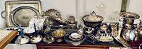 Wide shot showing the full range of silver and silver-plated items including trays, bowls, teapots, candle holders, and assorted utensils arranged on wooden furniture.