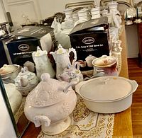 View of various tea pots, porcelain containers including a white embossed soup tureen with spoon and 7-piece soup and chili serve set in box on table with lace runner.