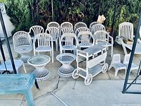 Wide view showing multiple white plastic patio chairs, spiral tables, bar cart, bench, and small children's chairs grouped outside.